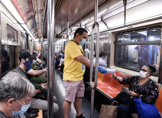 A volunteer in a yellow MTA mask force t-shirt stands in a subway car and gives outa mask to a woman, who is masked, sitting on a seat